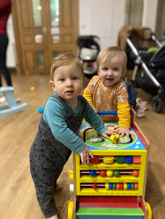 Two white, fair-haired toddlers leaning on a toy cube with abacus. Both are looking at the camera and grinning. 