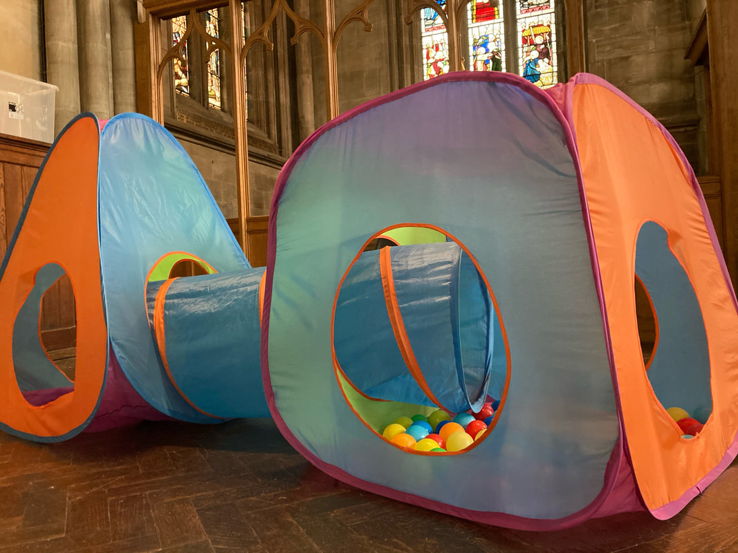 A ball pit cube, tunnel and pyramid set. In the background are a wooden screen and stained glass windows