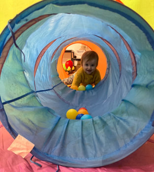 A fair haired white toddler grins from the far end of a blue fabric tunnel, with ball pit balls inside